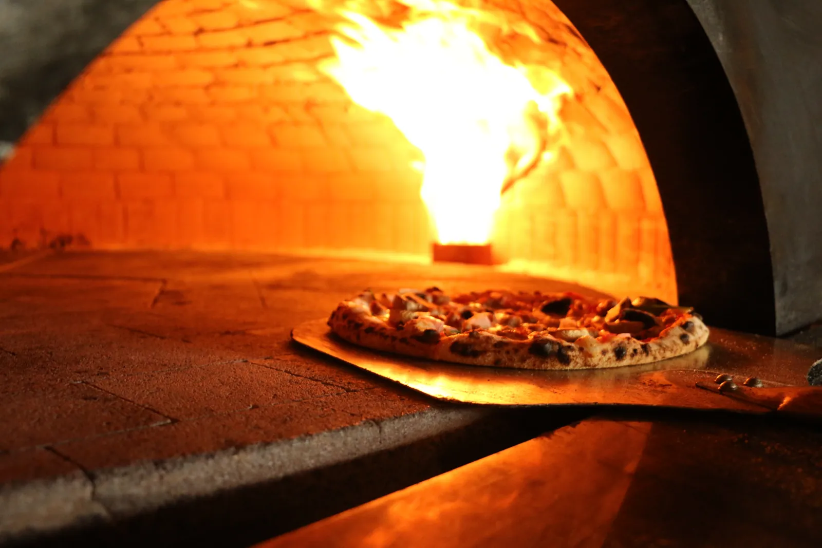 A pizza on a wooden peel entering the Longshot brick oven, with open flame against the oven wall