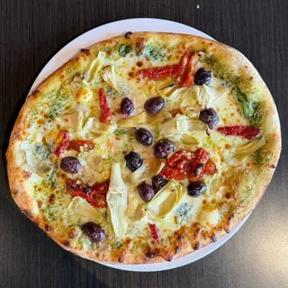 The Roasted Med, a Longshot specialty pizza seen from above: pesto Sauce, Mozzarella & Provolone, Spinach, Artichoke, Sun-Dried Tomato, Kalamata Olives, Feta Cheese, on a blistered brick-oven crust (vegetarian)