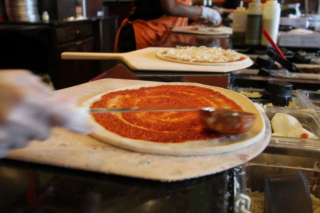 A gloved hand ladling house marinara onto a hand-stretched pizza base at the Longshot make line