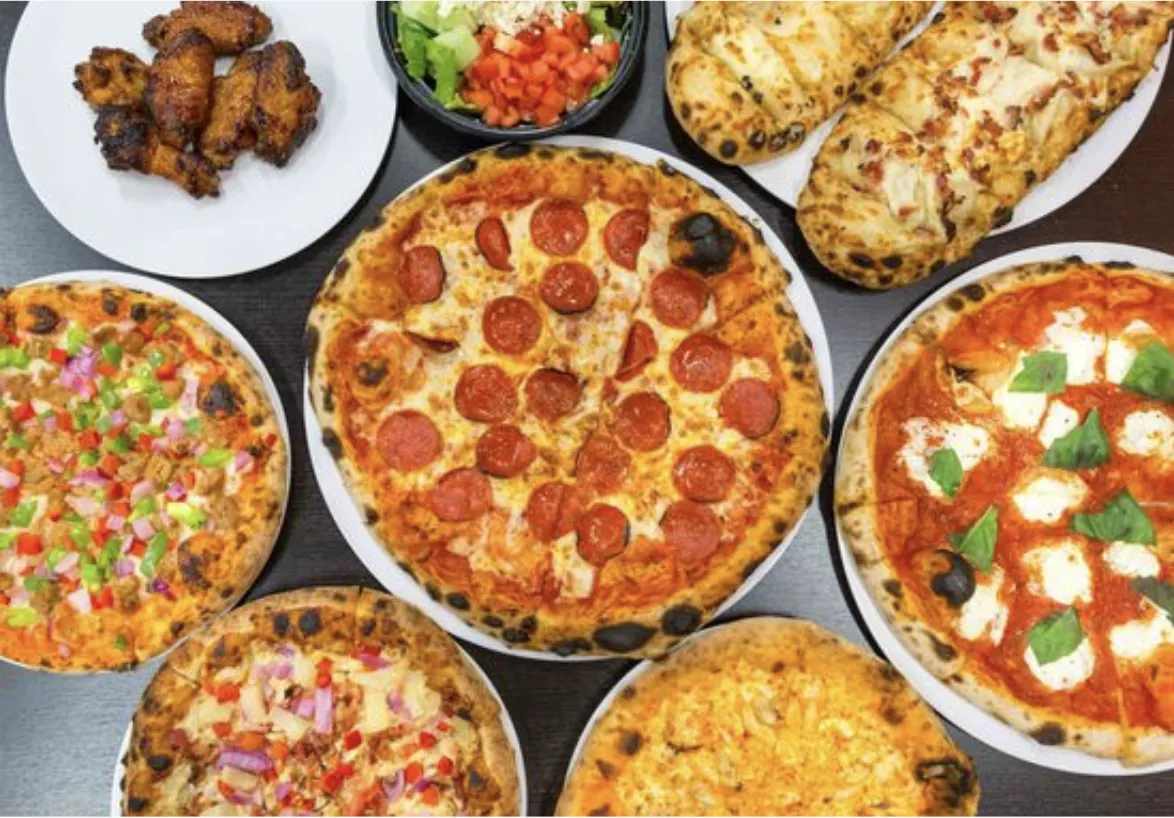 A Longshot catering spread: five pizzas, a tray of wings, a salad and cheesy bread laid out on a table