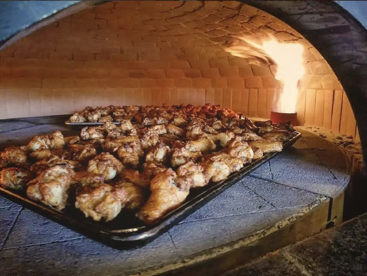 Trays of Longshot wings baking on the stone deck of the brick oven, flame visible at the back