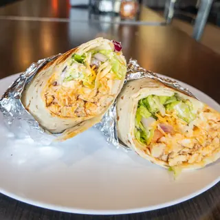 Buffalo Chicken Wrap, halved on a white plate so the filling shows: grilled Chicken, Cheddar, Romaine Lettuce, Tomato, Red Onion, Buffalo Sauce, Ranch Drizzle. Served warm in foil or cold.