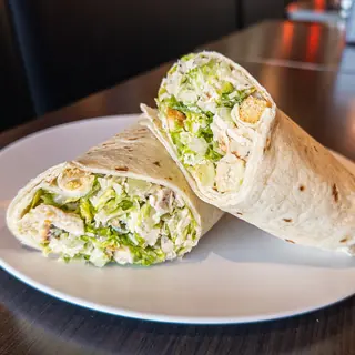 Chicken Caesar Salad Wrap, halved on a white plate so the filling shows: grilled Chicken, Asiago Cheese, Romaine Lettuce, Croutons, Caesar Dressing. Served cold.